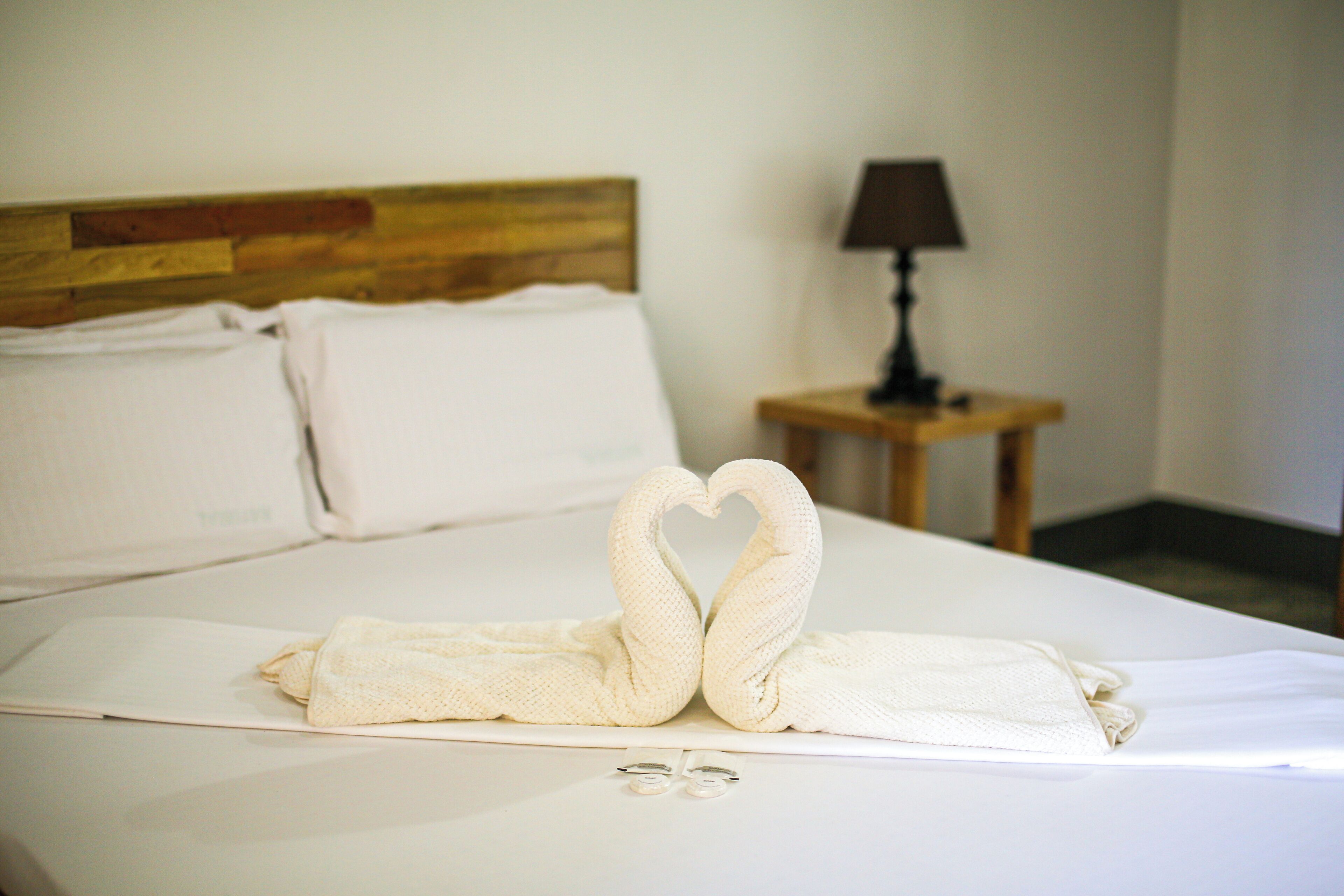 Una cama bien hecha con una toalla dispuesta en forma de corazón, un cabecero de madera y una mesita de noche con una lámpara.