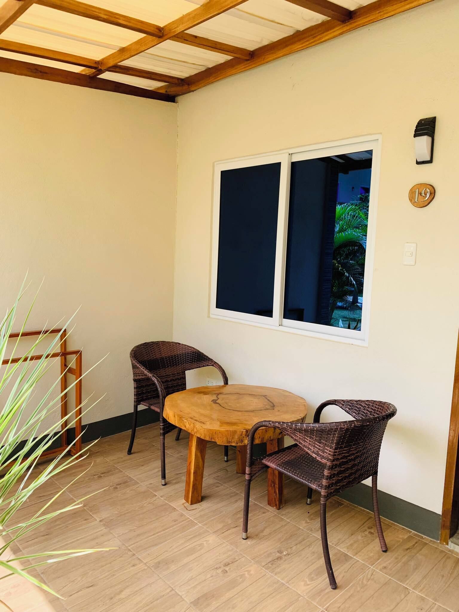 A small patio with a round wooden table and four wicker chairs. There is a window with a view of greenery outside.