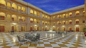 A spacious courtyard with checkered flooring and multiple tables and chairs, surrounded by arched windows and doors.