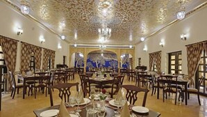 Een ruime eetruimte met een versierde plafond en kroonluchters, meerdere eettafels gedekt met witte tafelkleden en servetten, en een grote muurschildering op de achterwand.