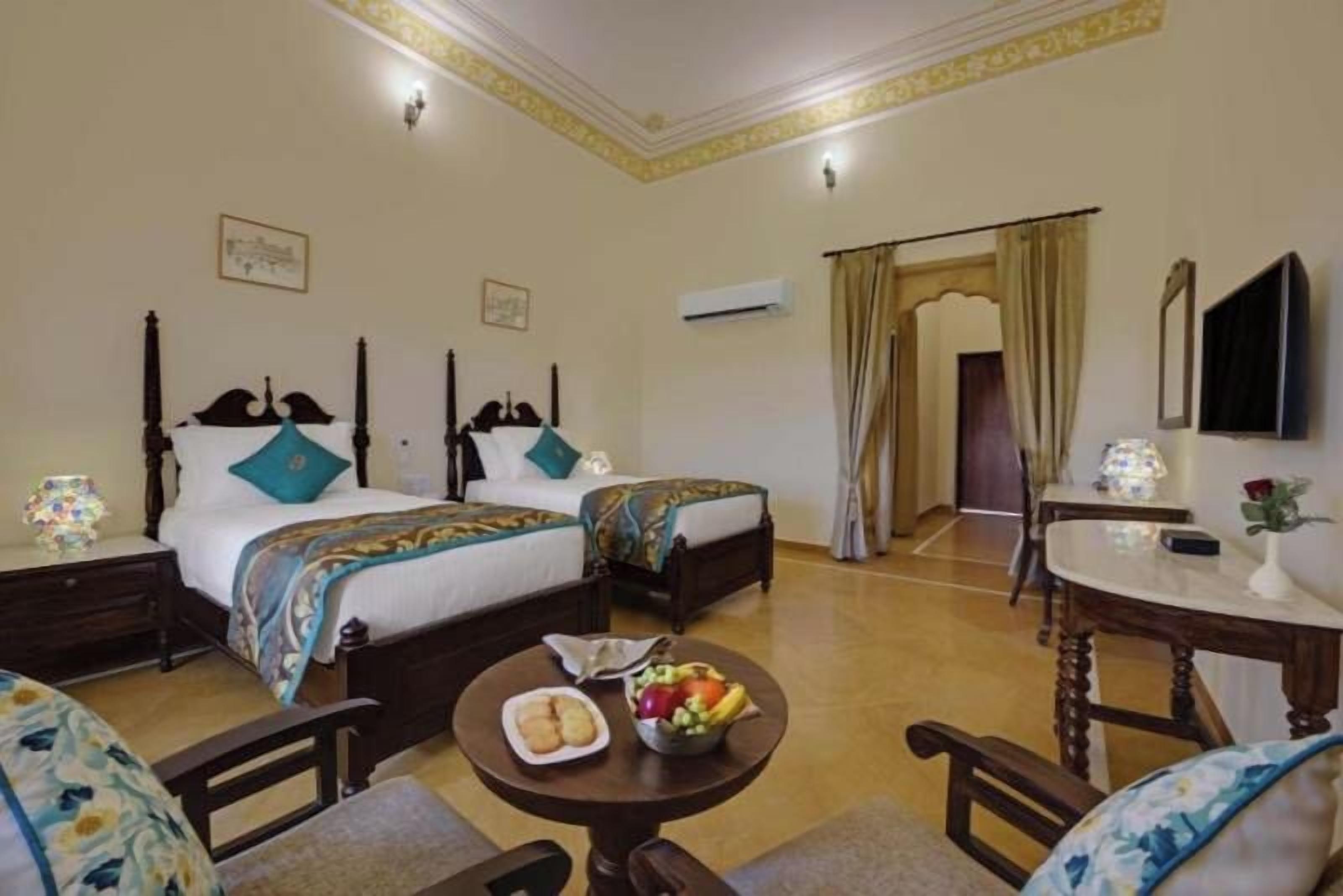 A hotel room with two beds, a dining table with fruit, a chair, and a small table with a lamp.