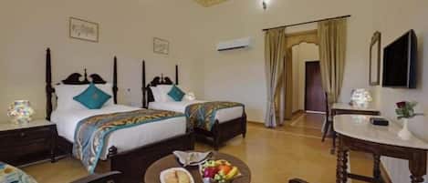 A hotel room with two beds, a dining table with fruit, a chair, and a small table with a lamp.