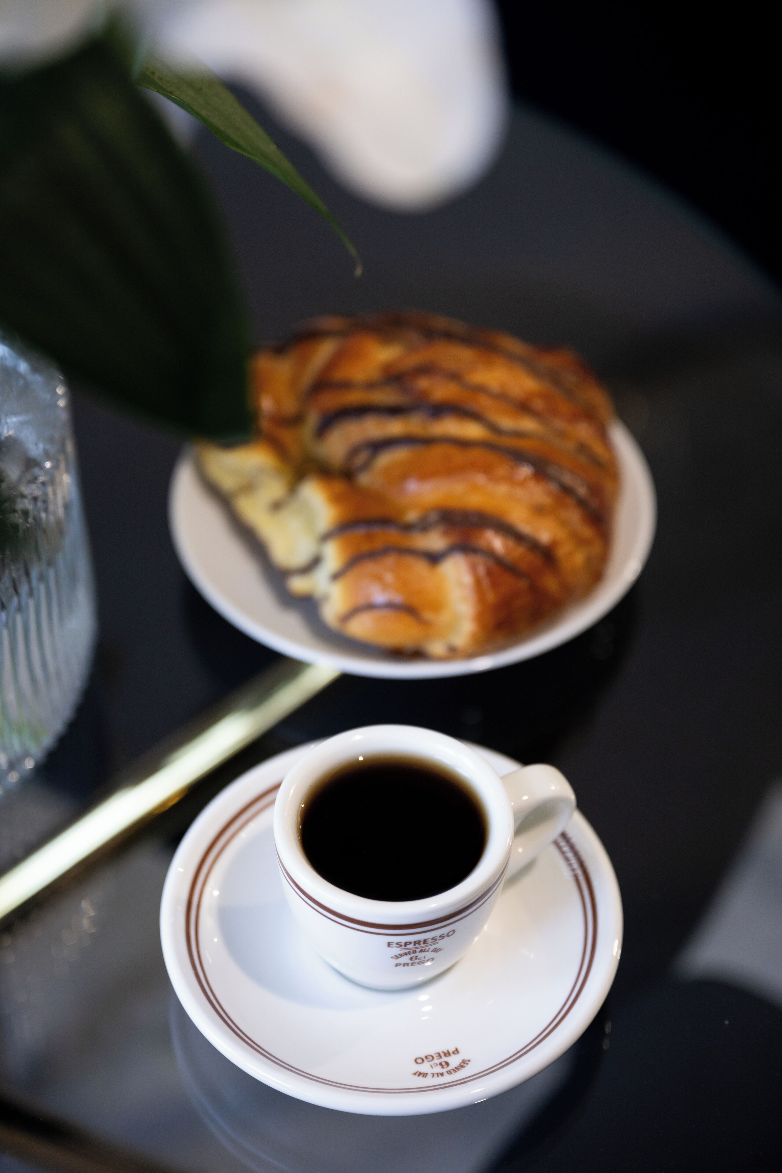 Una taza de espresso sobre un platito con un plato de croissants al fondo.