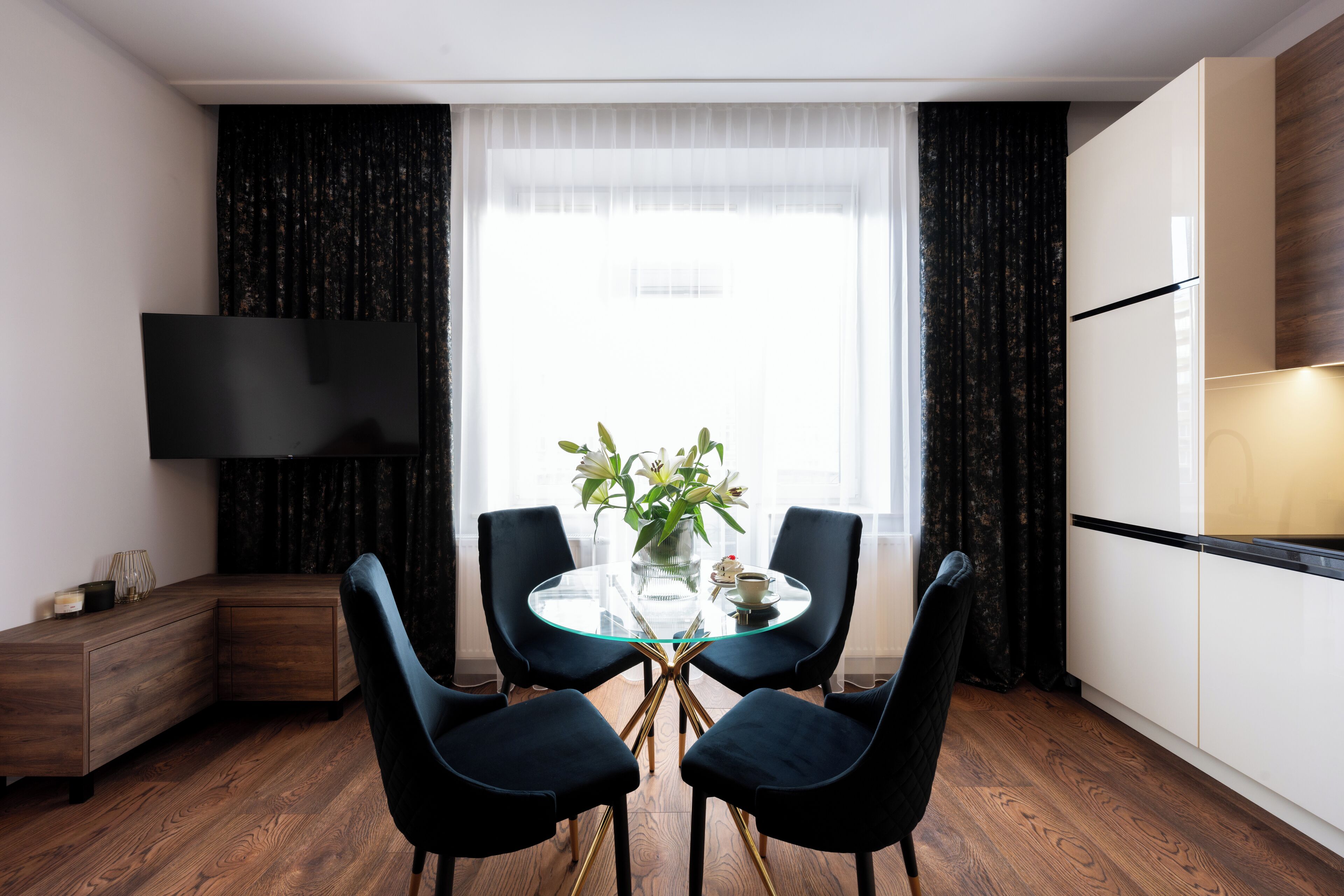 Un comedor moderno con una mesa de vidrio, cuatro sillas negras, un jarrón con flores y un televisor en la pared.