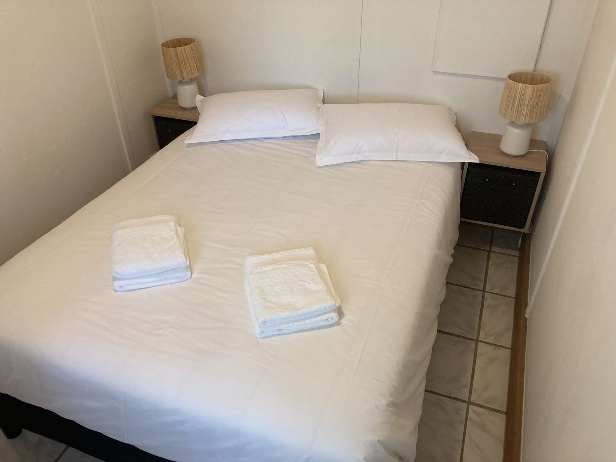 Una cama bien hecha con sábanas blancas, dos almohadas y dos toallas dobladas sobre la cama. Hay dos mesitas de noche con lámparas a cada lado de la cama.
