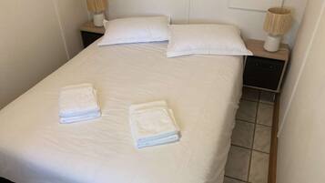 Una cama bien hecha con sábanas blancas, dos almohadas y dos toallas dobladas sobre la cama. Hay dos mesitas de noche con lámparas a cada lado de la cama.