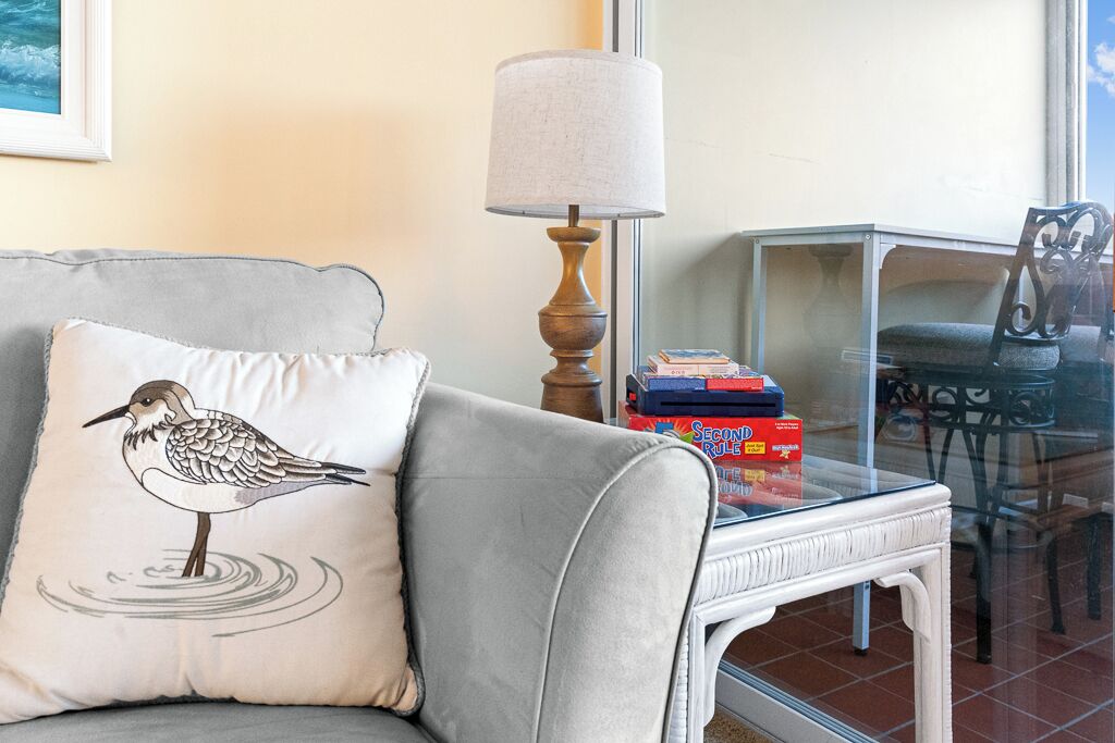 A living room with a grey sofa, a white pillow with a bird design, a table with a lamp, and a glass table with books and a chair.