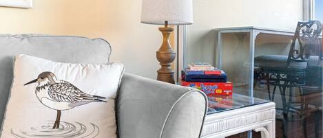 A living room with a grey sofa, a white pillow with a bird design, a table with a lamp, and a glass table with books and a chair.