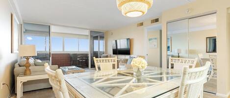 A spacious room with a glass-top dining table surrounded by white chairs, a woven pendant light, and a flat-screen TV mounted on the wall.
