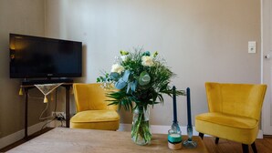 Un salón con un televisor de pantalla plana, dos sillones amarillos, una mesa de centro de madera y un florero con flores.