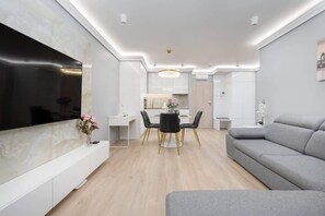 Una sala moderna con un televisor de pantalla plana, un sofá gris, un comedor con una mesa redonda y una cocina al fondo.