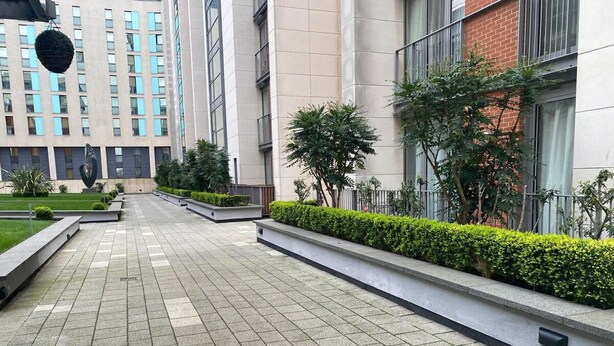 A modern building with a tiled walkway, green hedges, and a hanging planter. - Royal Docks Luxury 3 Bedroom Apartment (London)