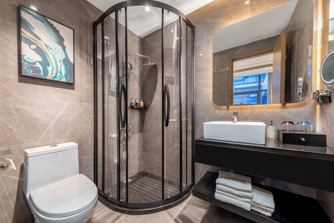 Un baño moderno con ducha de vidrio, inodoro blanco, tocador negro con espejo y pared con azulejos de mármol.