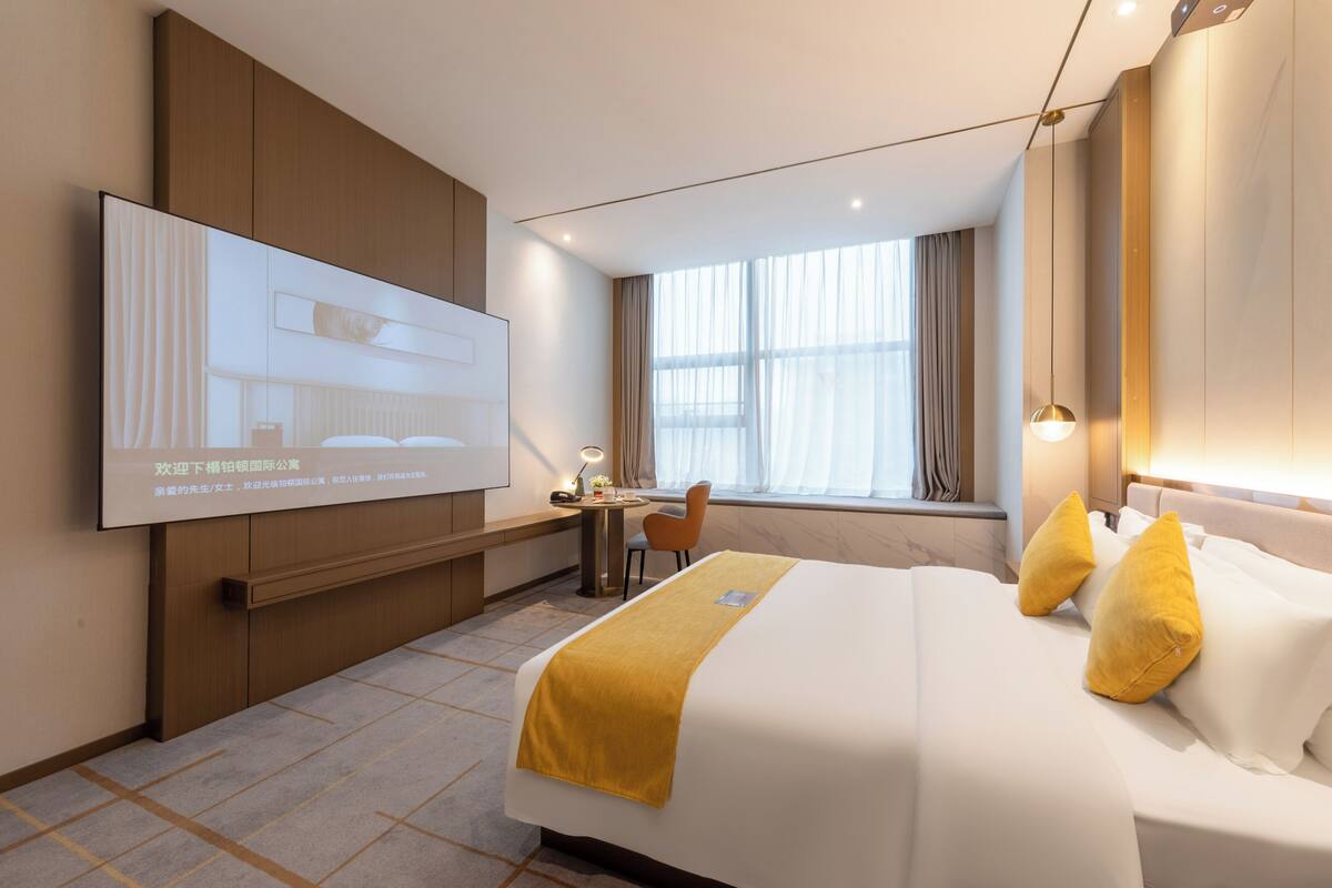 A modern hotel room with a large bed, a desk, a chair, and a TV mounted on the wall.