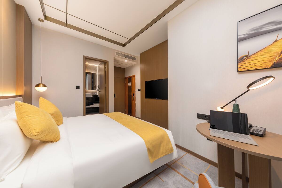 A hotel room with a bed, a desk with a laptop, a TV, and a painting on the wall.
