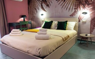 Un letto rifatto con lenzuola bianche e cuscini verdi, un comodino con una lampada e una sedia in una stanza con un murale tropicale sulla parete.