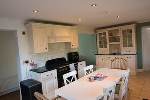 A compact kitchen with white cabinets, a black stove, and a dining table with white chairs. - Mumbles - House - Seaside - Sleeps11 - Petfriendly (Swansea)