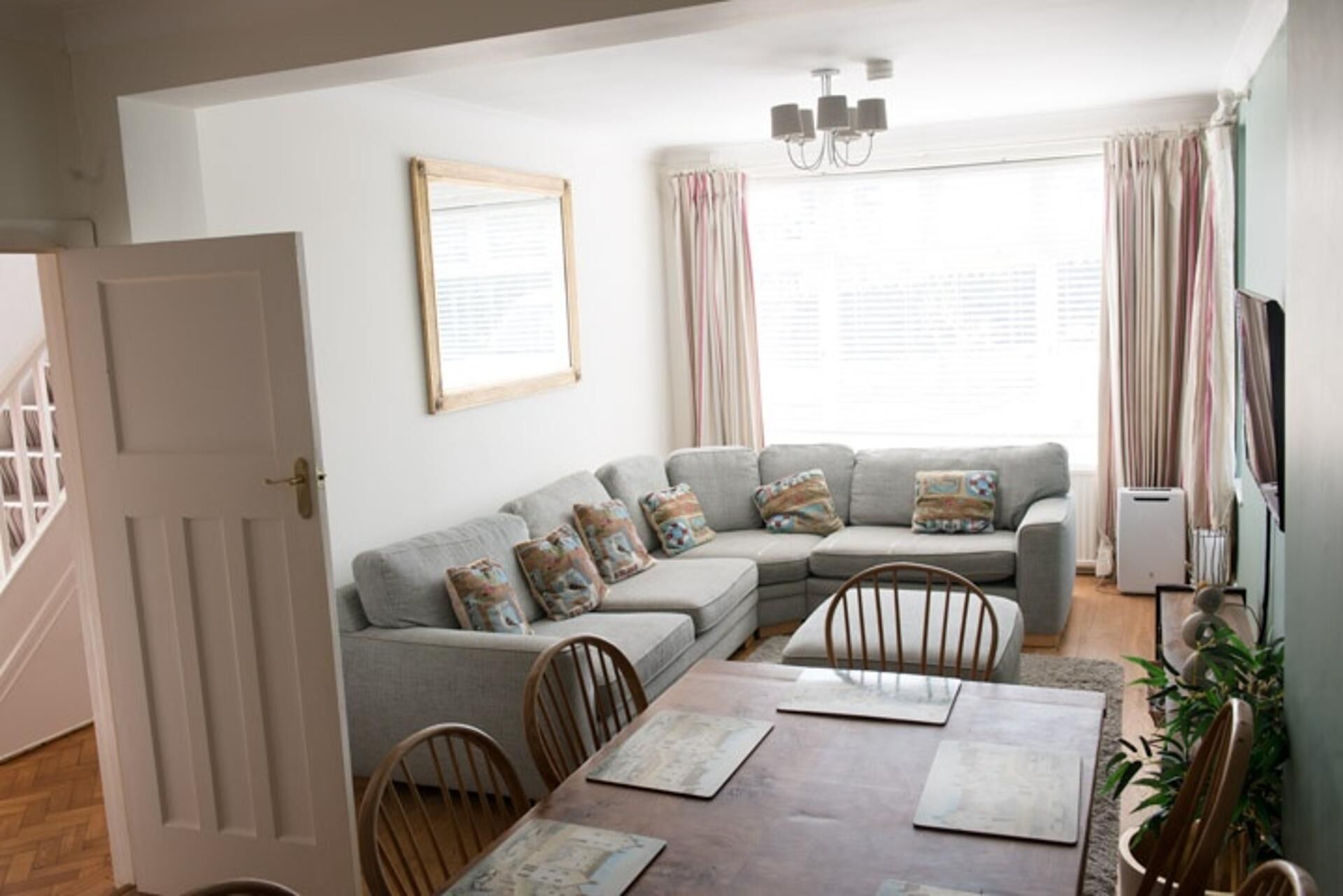 Una sala de estar con un sofá seccional gris, sillas de madera, una mesa de comedor con individuales, una araña y un ventanal con cortinas.