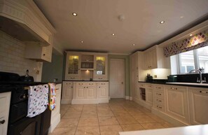 A kitchen with white cabinets, a tiled floor, and a window with a patterned curtain. - Mumbles - House - Seaside - Sleeps11 - Petfriendly (Swansea)