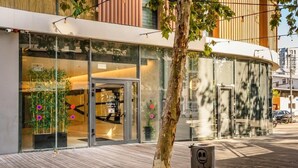 A modern building with a curved facade, large glass windows, and a wooden deck area with a tree and trash bin. - Luxury 3BR/2BA Apartment with Terrace in TLV | By SWEETYGUEST (Tel Aviv-Yafo)