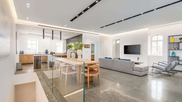 Ein modernes Wohnzimmer mit Glastrennwand, einem Essbereich mit Holzmöbeln, einem grauen Sofa und einem an der Wand montierten Fernseher.