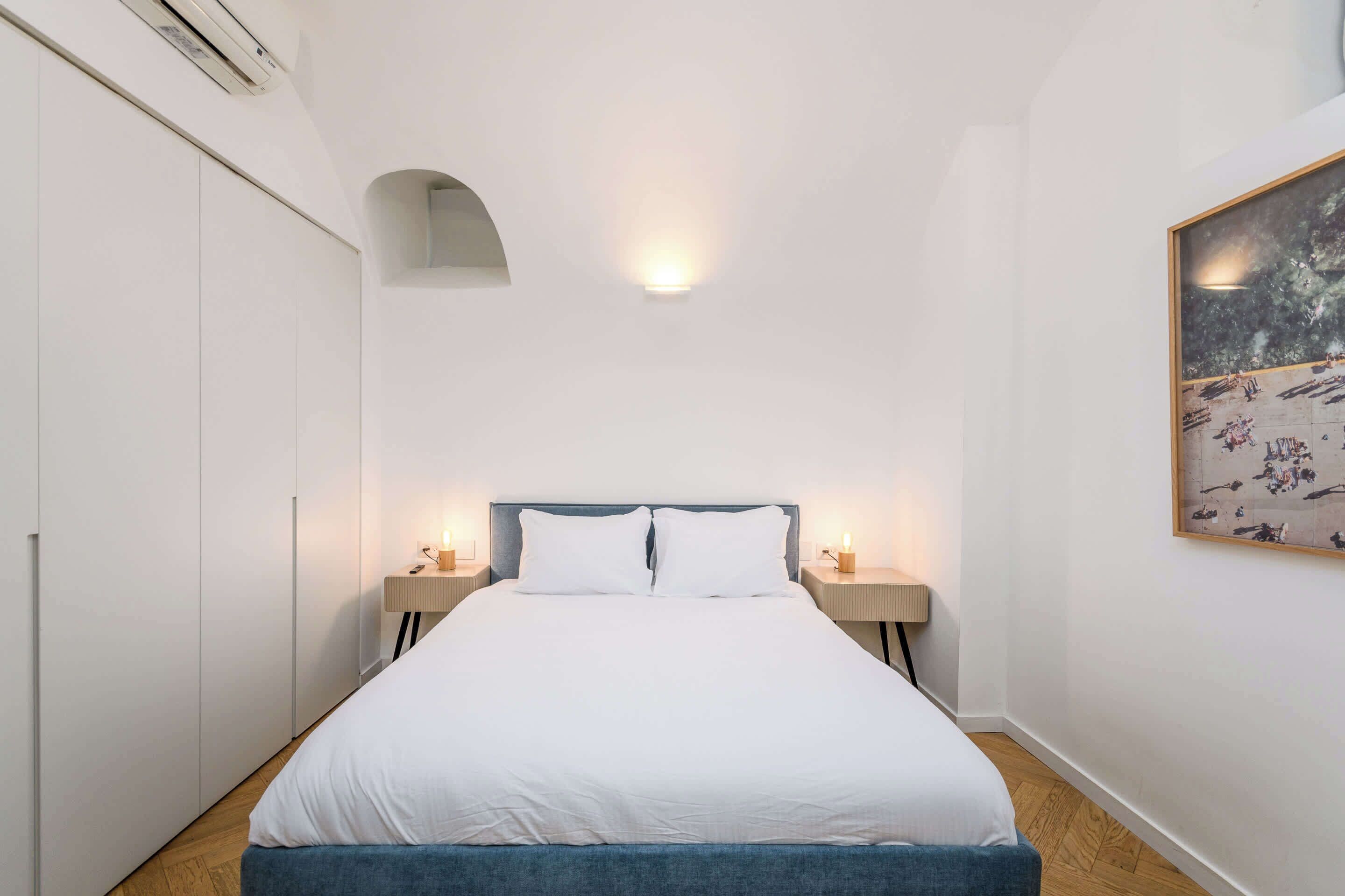 Une chambre à coucher comprenant un lit, deux tables de chevet, une lampe fixée au mur, un tableau et une armoire.