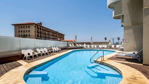 Una piscina en la azotea con agua azul cristalina, rodeada de sillas reclinables blancas y una terraza de madera.