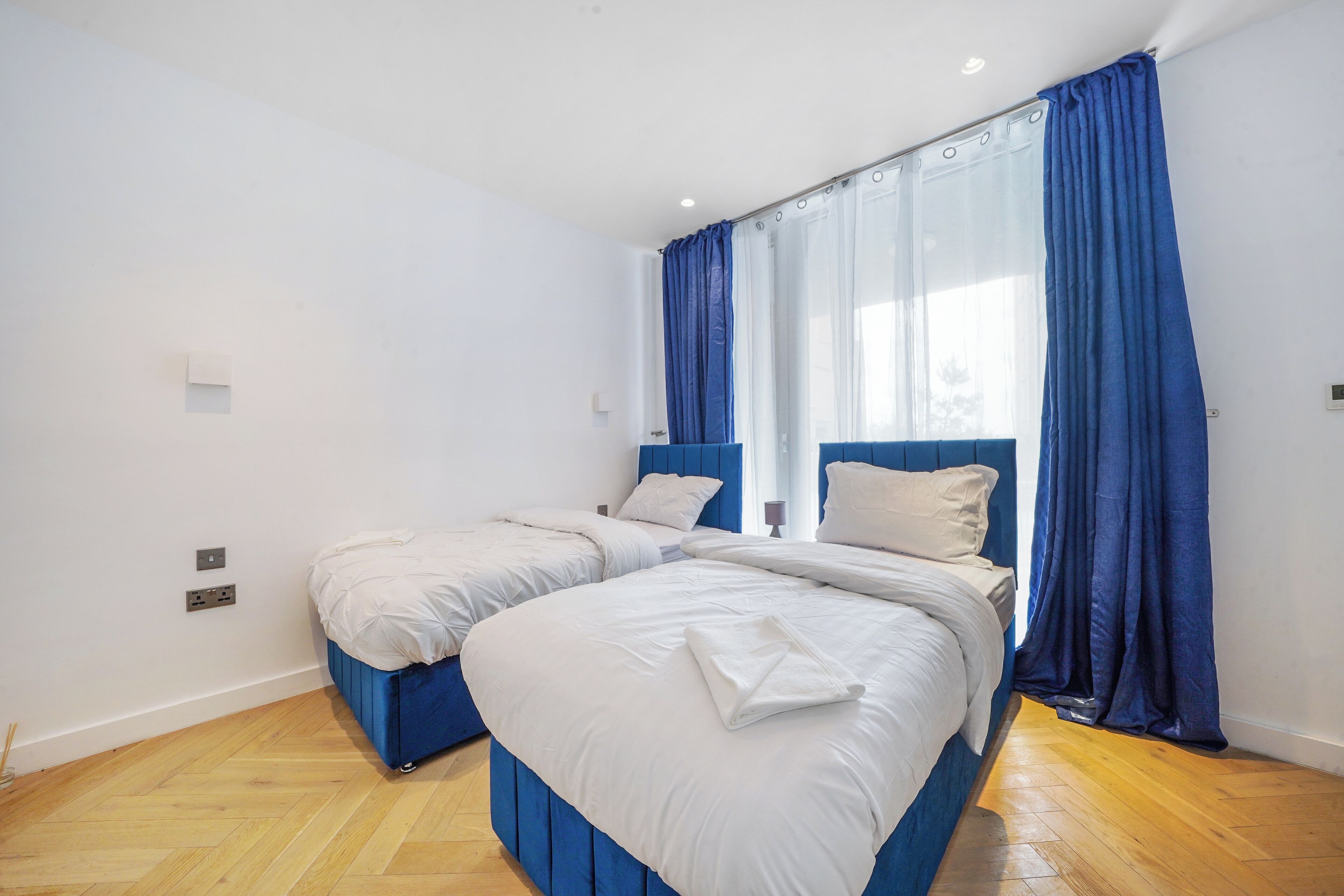 A double room with two beds, a large window with blue curtains, and a wooden floor.