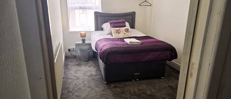 A bedroom with a bed, a bedside table, a lamp, a window, and a wall-mounted shelf.