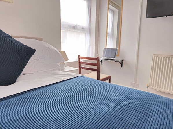 A neatly made bed with a blue bedspread, a wooden chair, a laptop on a small table, and a radiator. - PRIVATE ROOMs In TOWN CENTRE FREE PARKING WIFI AND PATIO WITH AA STAY LTD (Aylesbury)