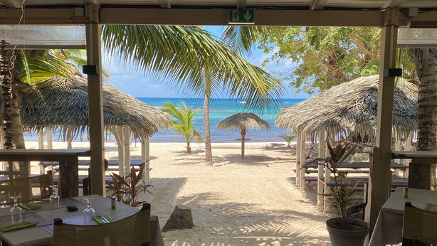 Un restaurant situé en bord de mer, doté de parasols en chaume, de tables et de chaises en bois, et offrant une vue sur une plage de sable et l’océan.