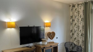 Un salon doté d’un téléviseur à écran plat, d’un meuble de rangement en bois, de deux lampes fixées au mur, d’un rideau à motifs et d’un tapis vert.