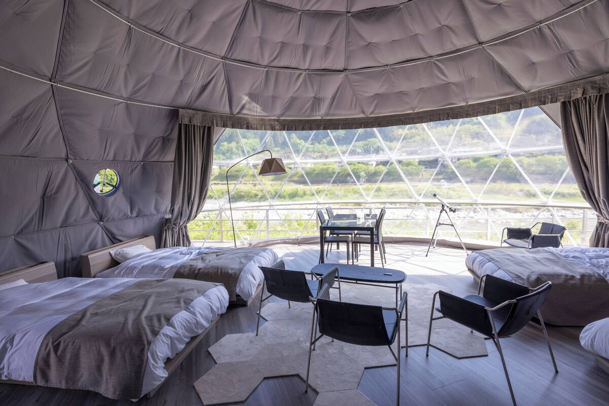 A tented room with two beds, a table, chairs, and a view of a greenhouse structure.