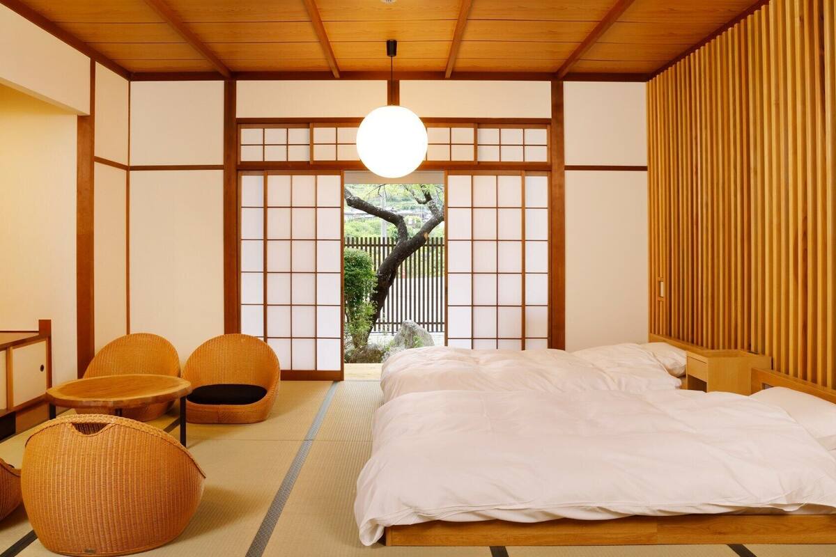 A traditional Japanese-style bedroom with tatami flooring, a low wooden bed, and a view of a tree outside.