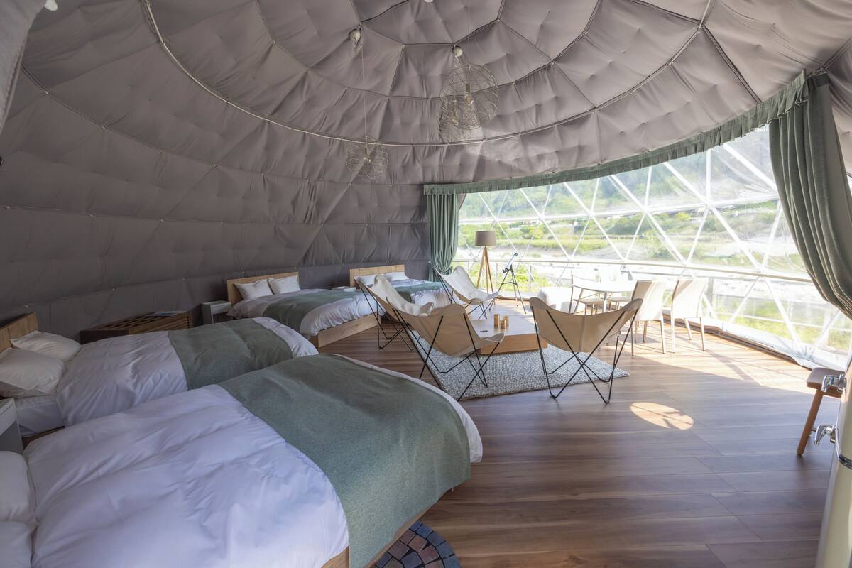 A tented accommodation with two beds, a dining area, and a view of greenery through large windows.