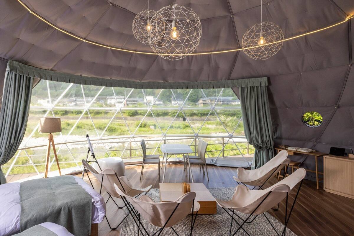 A tent-style room with a bed, chairs, a table, and a view of a field through large windows.