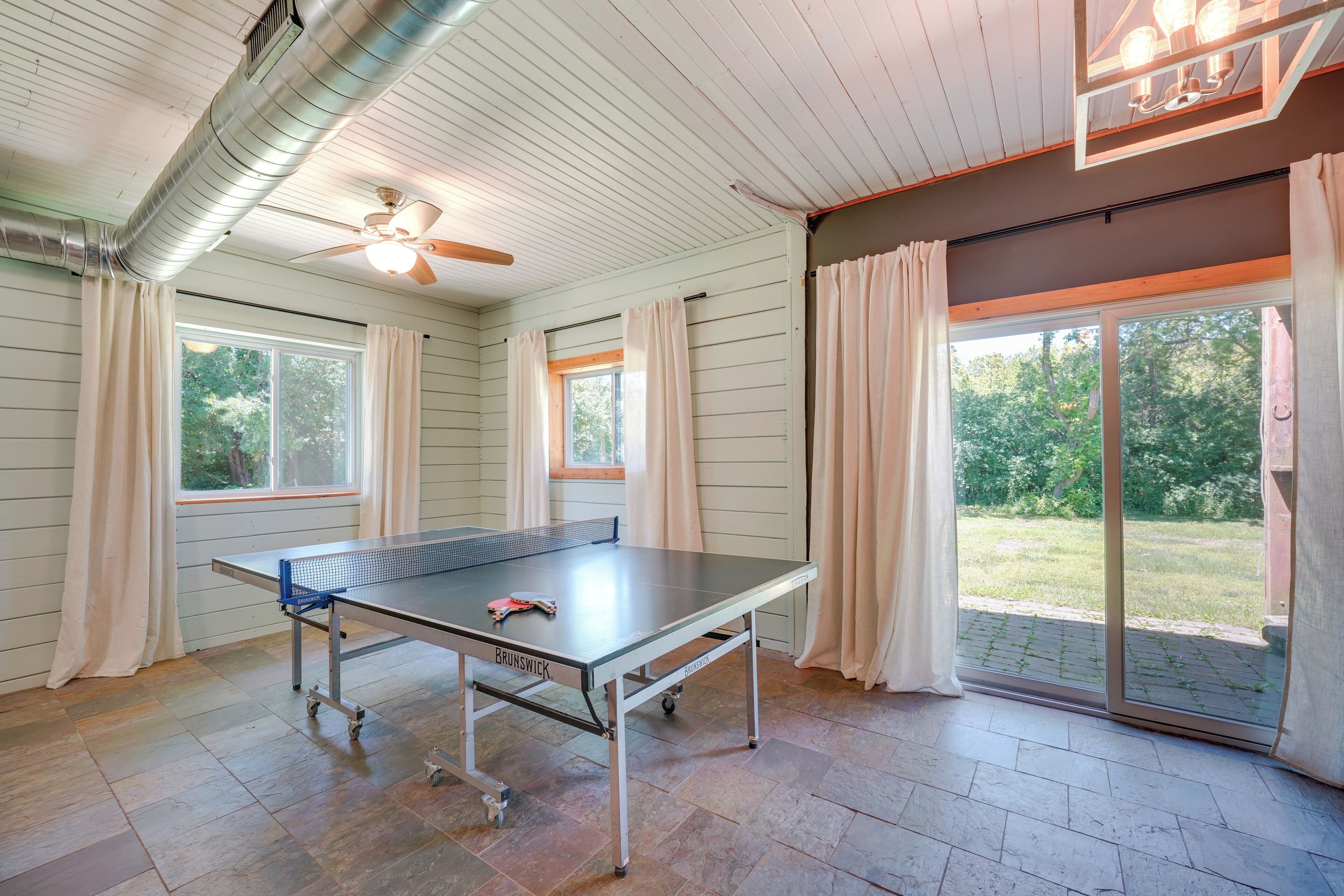 A ping pong table in a room with wooden walls, a ceiling fan, and large windows with curtains.