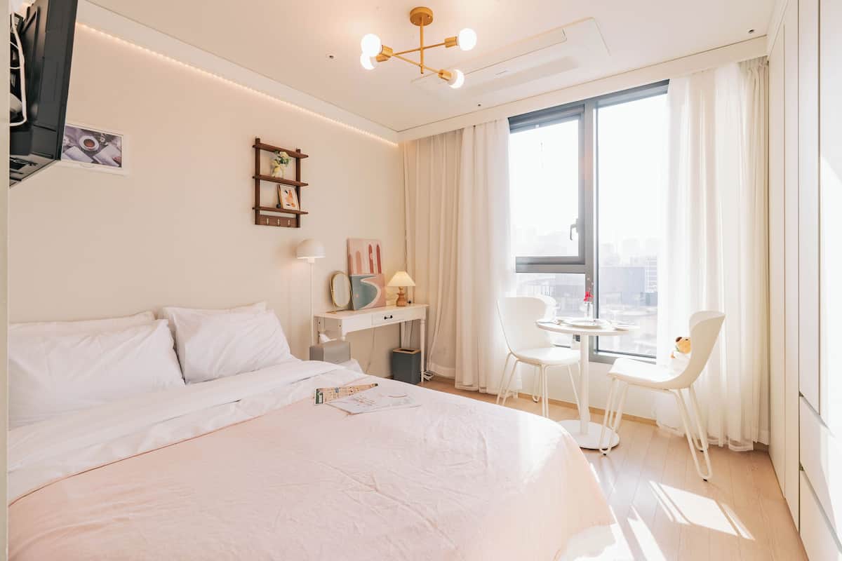 A hotel room with a large bed, a desk with a chair, a small table, and a view of the city through the window.