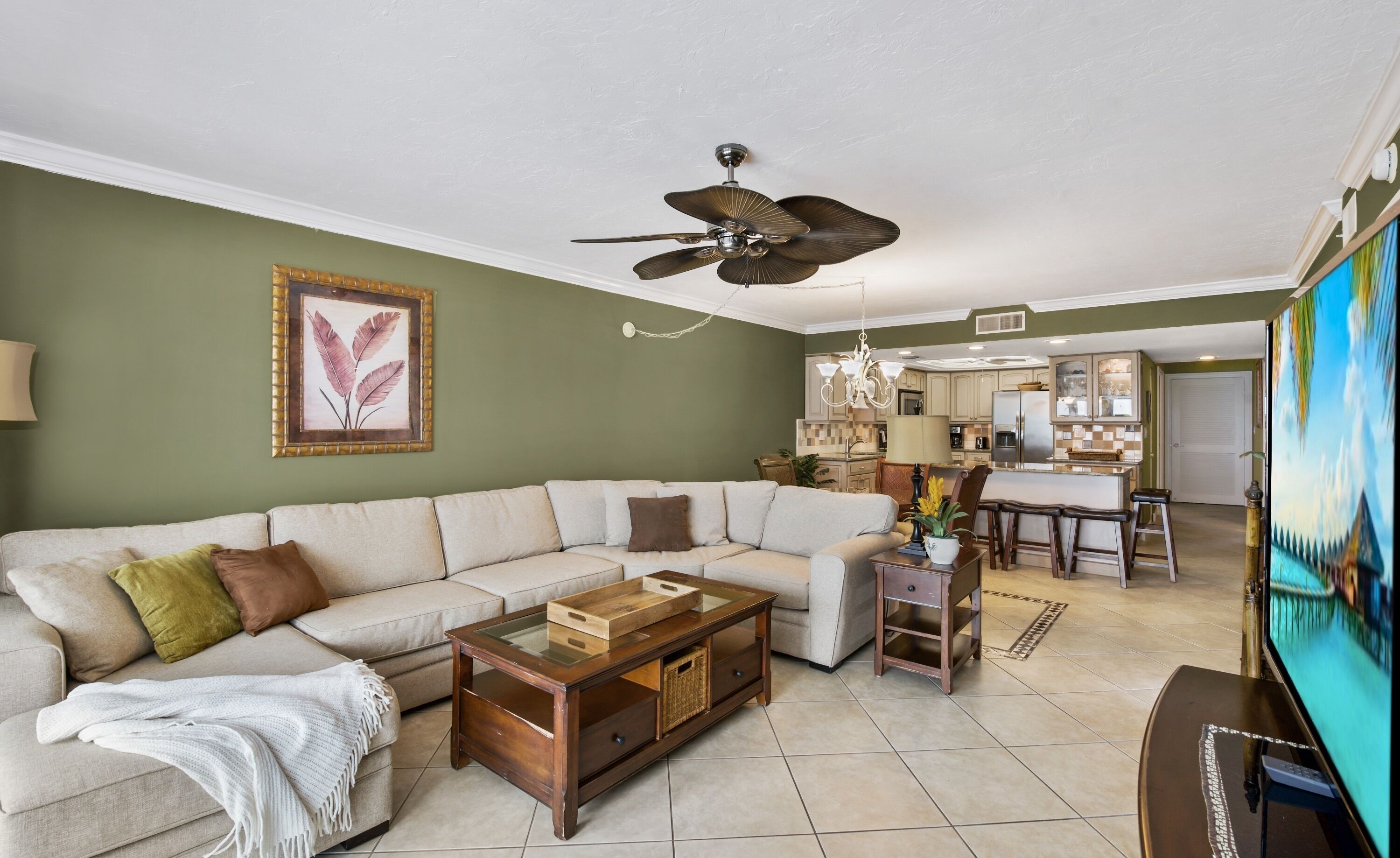 A living room with a sectional sofa, a coffee table, and a TV. There is a ceiling fan, a chandelier, and a kitchen in the background.