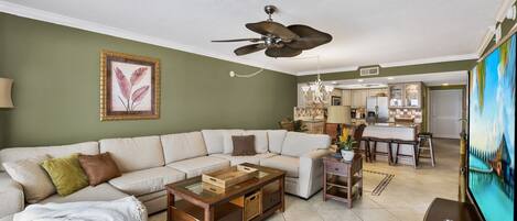 A living room with a sectional sofa, a coffee table, and a TV. There is a ceiling fan, a chandelier, and a kitchen in the background.