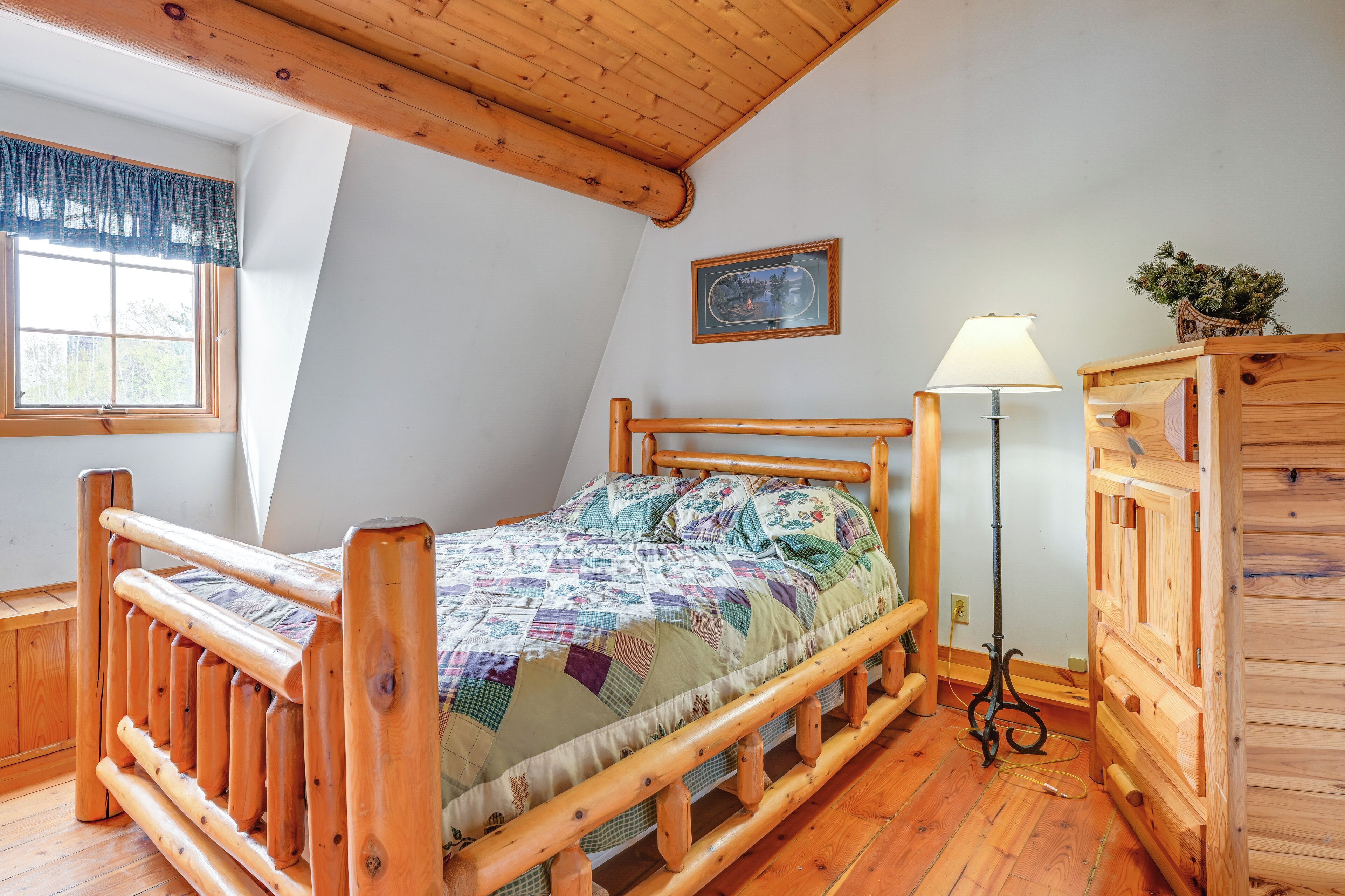 A wooden bed with a quilt, a wooden dresser, a lamp, and a framed picture on the wall.