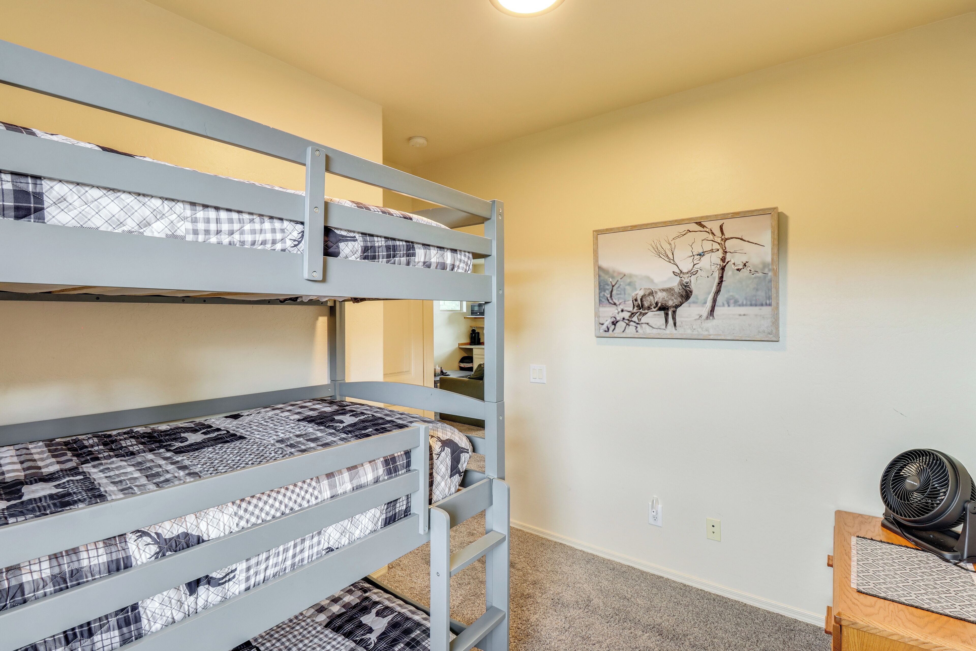 A bunk bed room with a checkered bedspread, a framed picture of deer, and a fan on a wooden table.