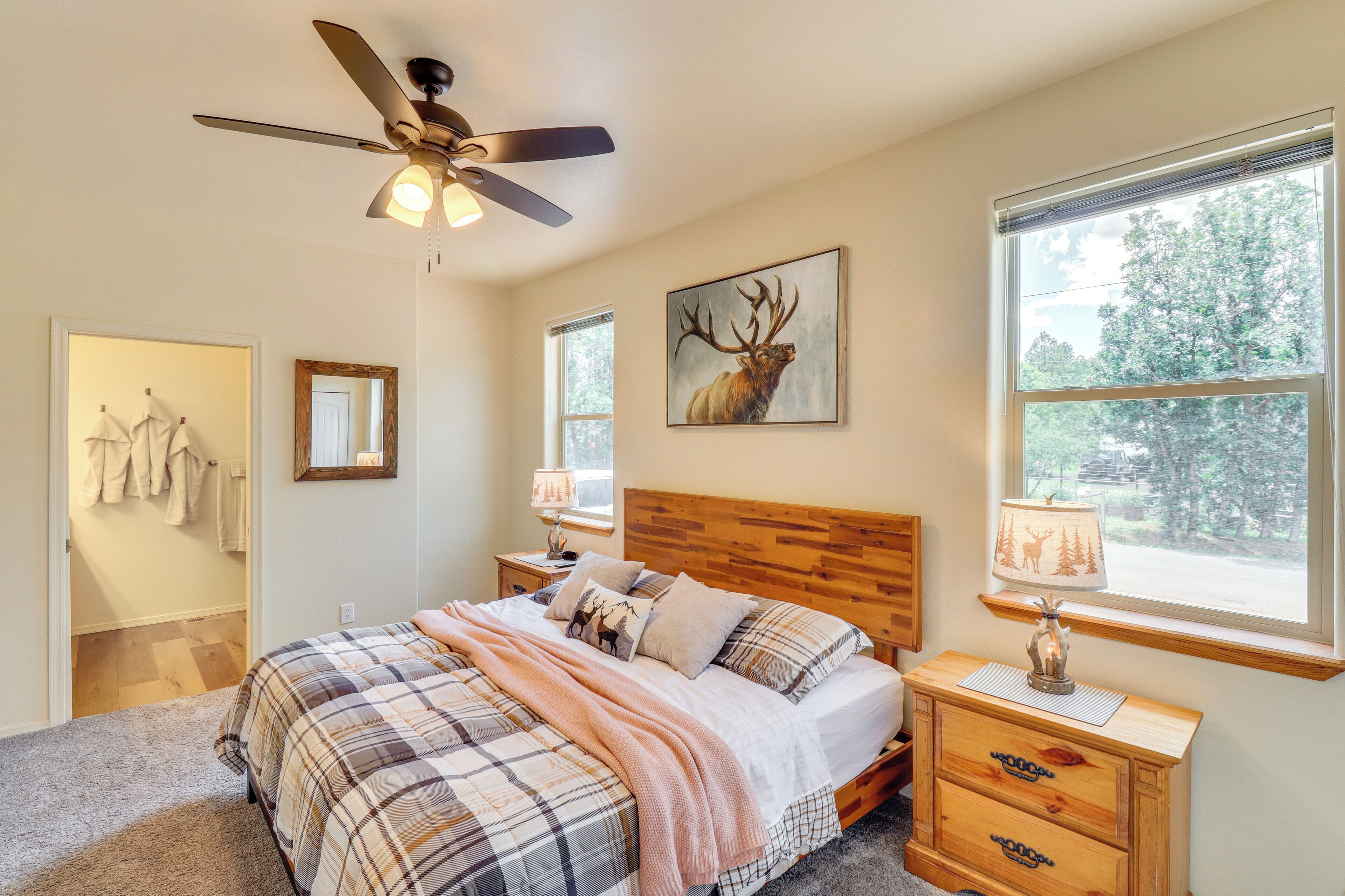 A bedroom with a wooden headboard, a ceiling fan, a large window with a view of trees, a bed with plaid bedding, and a nightstand with a lamp.