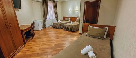 Habitación de hotel con piso de madera, una cama con toalla enrollada, un escritorio, un refrigerador pequeño y un televisor montado en la pared.