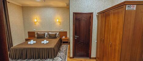 A hotel room with a bed, two bedside lamps, a wooden wardrobe, and a door with a 'No Smoking' sign.
