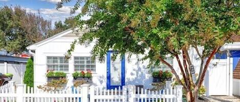Ein weißes Haus mit einer blauen Tür, umgeben von einem weißen Zaun und einem Baum davor.