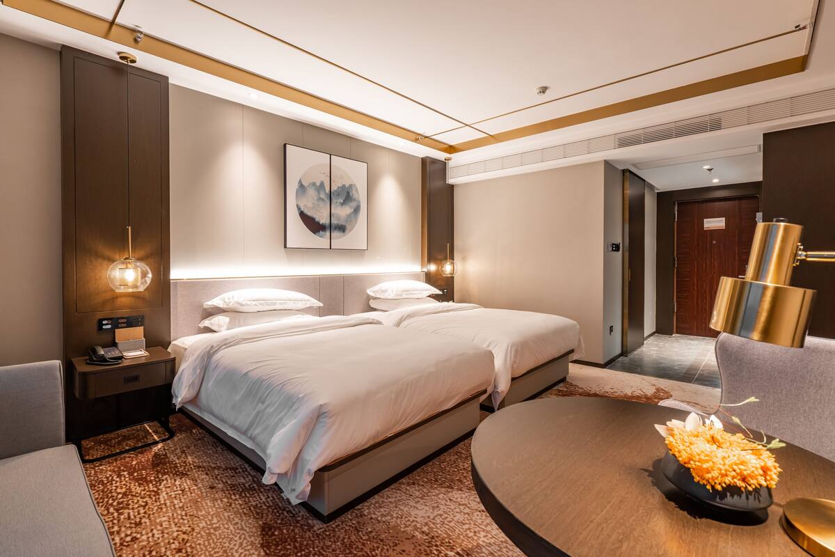 A modern hotel room with a large bed, bedside tables, a round coffee table with flowers, and a painting on the wall.