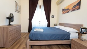 A neatly arranged bedroom with a bed, bedside tables, a fan, and a painting on the wall.