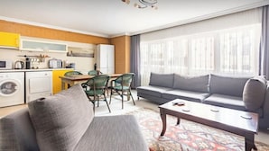 Una sala de estar con sofá, una mesa de centro y un comedor con sillas y una mesa. Hay una zona de cocina con electrodomésticos y una ventana con cortinas.