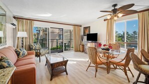 A spacious living room with a sofa, a glass coffee table, wicker chairs, a TV, and a balcony with a view of palm trees. - Pono Kai Resort J201, 2nd Floor, Walk to Beach (Kapaa)
